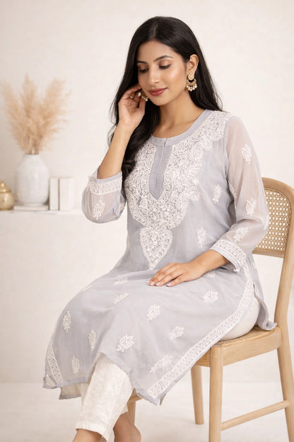 A woman seated on a wooden chair wearing a light gray embroidered kurta with white lace detailing and white pants, photographed in a softly styled indoor setting.