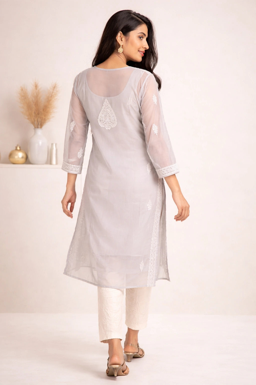 A woman modeling a light gray three-quarter sleeve kurta with white embroidery, paired with off-white pants and brown sandals, shown from a rear three-quarter view in a bright studio setting.