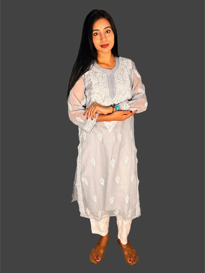 A woman modeling a light gray embroidered kurta with matching white pants and brown sandals, set against a solid dark gray background.