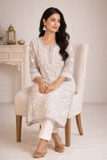 A woman sits on a cream upholstered chair wearing a light beige embroidered kurta with white pants and beige sandals in a softly lit indoor setting.