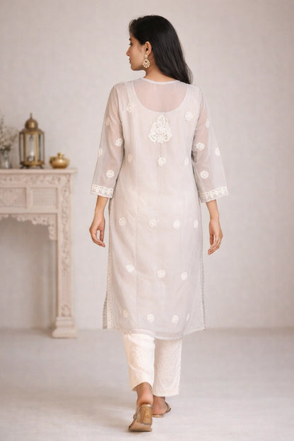 A woman wearing a light beige/ivory kurta with white floral embroidery and white pants, shown from the back in a softly lit indoor setting.