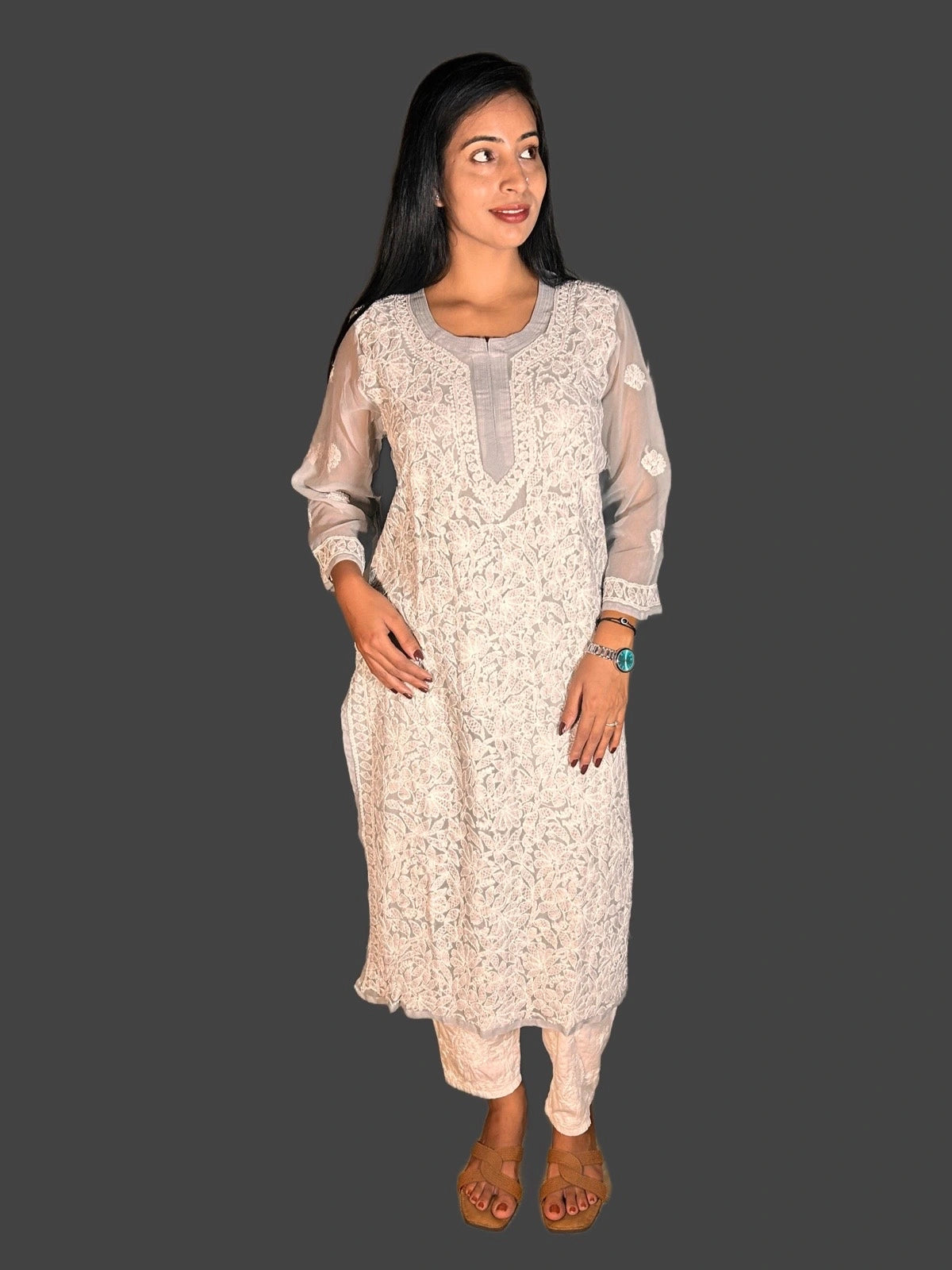 A woman modeling a traditional Indian outfit consisting of a cream-colored embroidered kurta with sheer sleeves and matching trousers, set against a solid dark gray background.