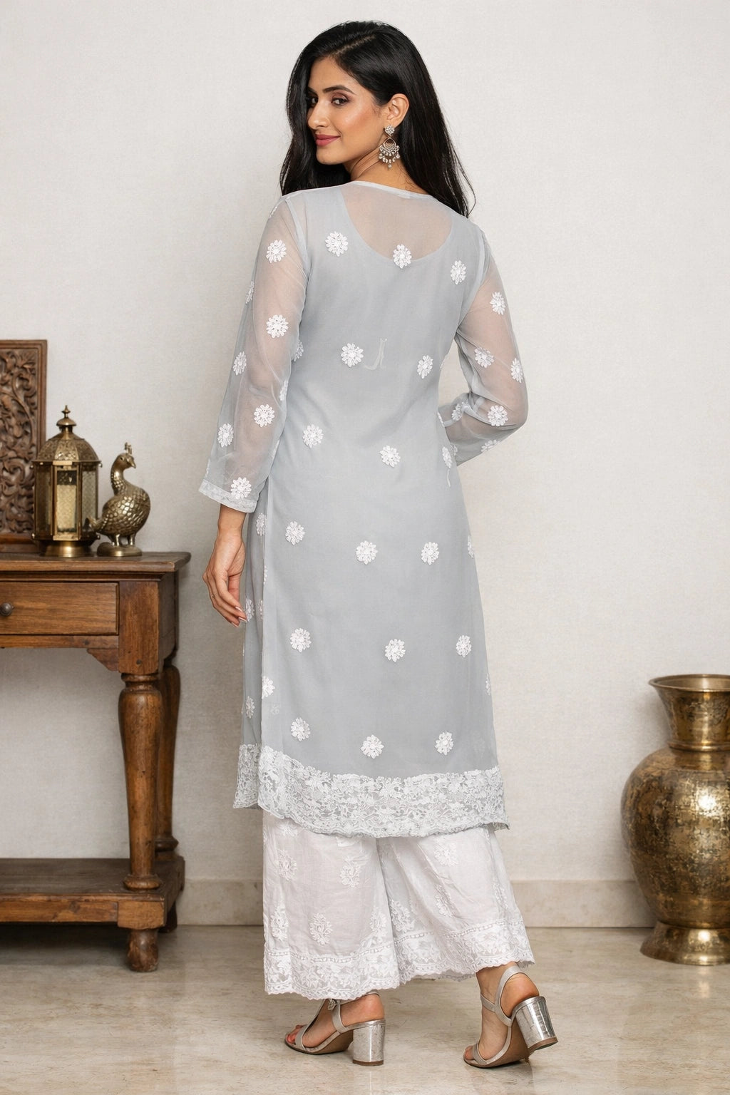 Woman wearing a lucknowi chikankari   light gray traditional outfit with white polka dots and lace details, standing in a room with wooden furniture and brass vases.