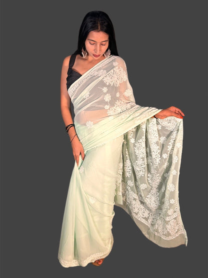 A woman modeling a pale mint green saree with white floral embroidery, paired with a black sleeveless blouse, set against a solid dark gray background.