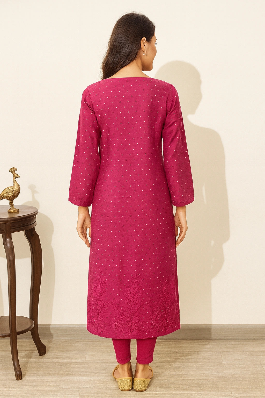 A woman viewed from the back wearing a magenta pink kurta with small white embroidered motifs, paired with matching pink pants and beige shoes, standing in a room with a light beige wall and wooden floor.