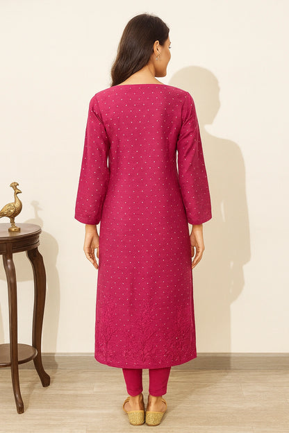A woman viewed from the back wearing a magenta pink kurta with small white embroidered motifs, paired with matching pink pants and beige shoes, standing in a room with a light beige wall and wooden floor.