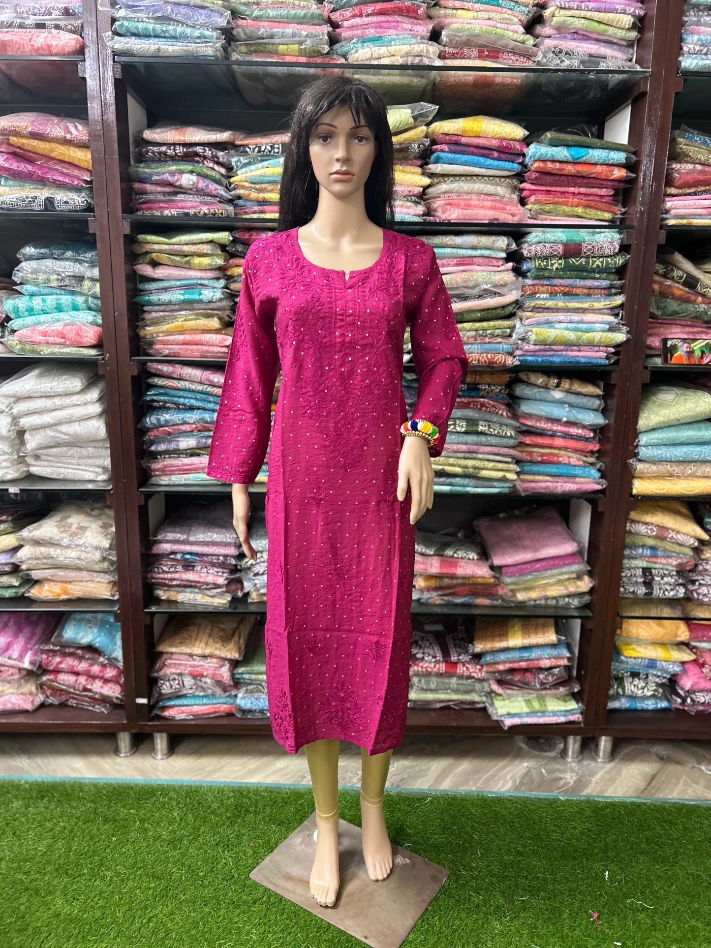 A mannequin wearing a bright magenta pink embroidered kurta dress, standing in front of shelves filled with folded colorful garments in a retail store.