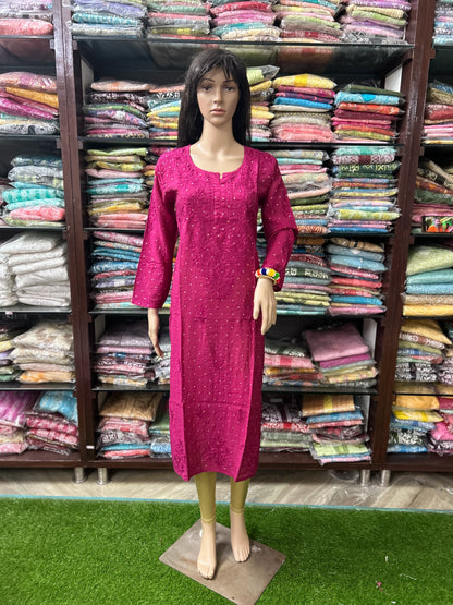 A mannequin wearing a bright magenta pink embroidered kurta dress, standing in front of shelves filled with folded colorful garments in a retail store.