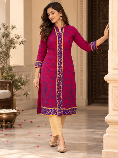 A woman models a magenta pink kurta with purple and gold embroidery, paired with beige trousers, in an elegant indoor setting with classical columns.