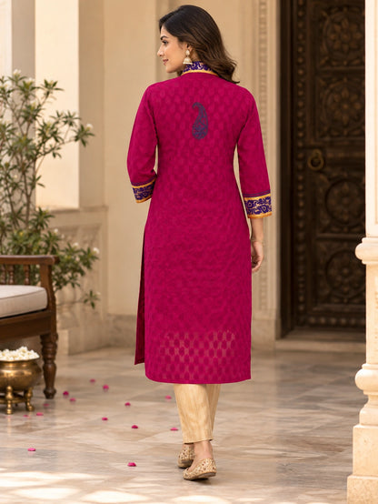 A woman wearing a bright magenta-pink kurta with a subtle geometric pattern, gold zari embroidery on the collar and cuffs, and beige trousers, shown from the back in an elegant outdoor courtyard setting.