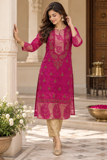 A woman models a bright magenta pink embroidered kurta with gold detailing, paired with beige trousers, in an elegant outdoor courtyard setting.