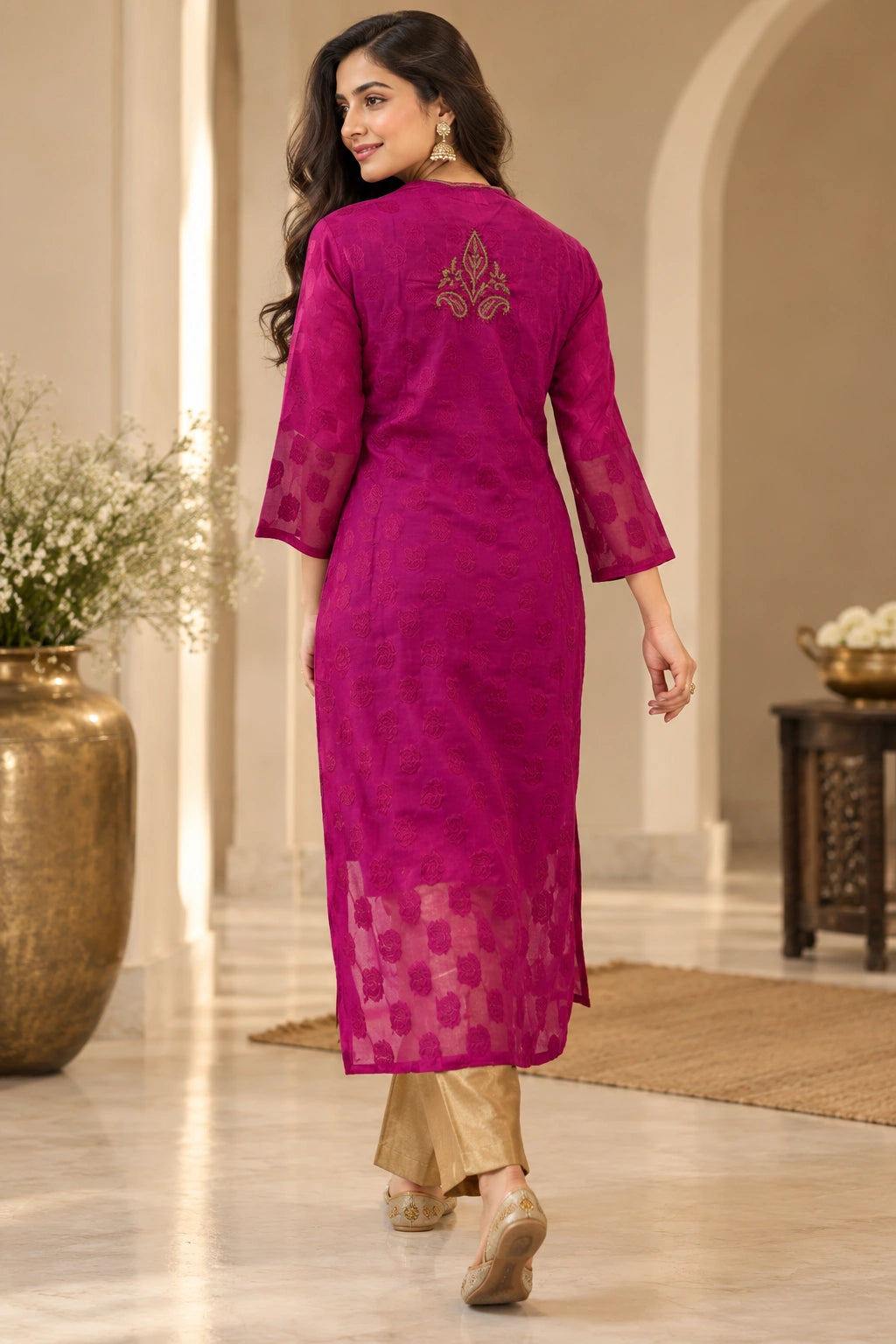 A woman wearing a magenta pink embroidered kurta with sheer sleeves and a checkered pattern near the hem, paired with beige trousers, shown from the back in an elegant indoor setting.