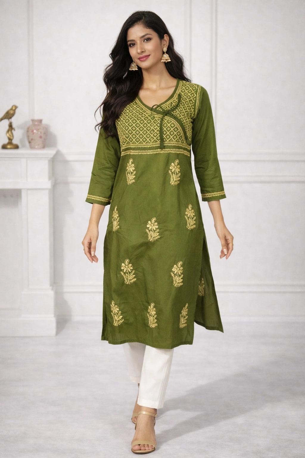 A woman models an olive green Indian kurta with gold embroidery, paired with white pants and beige sandals, in a bright indoor setting.