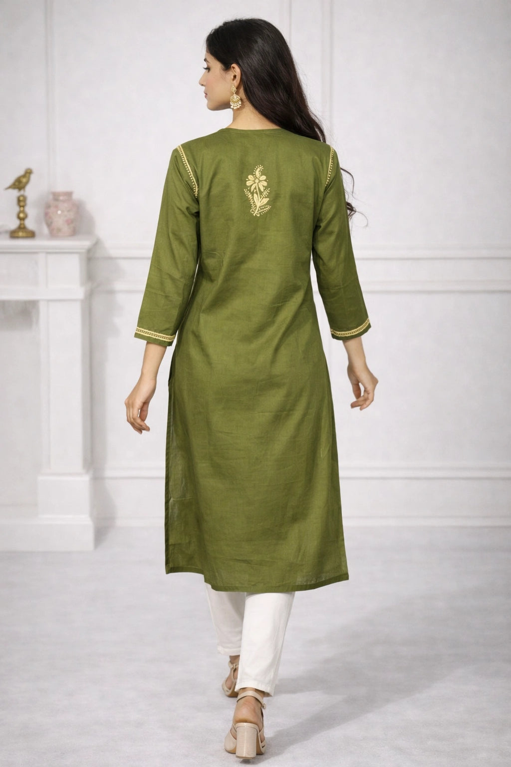 A woman shown from the back wearing an olive green kurta with gold floral embroidery on the upper back and gold-trimmed cuffs, paired with white pants in a bright indoor setting.