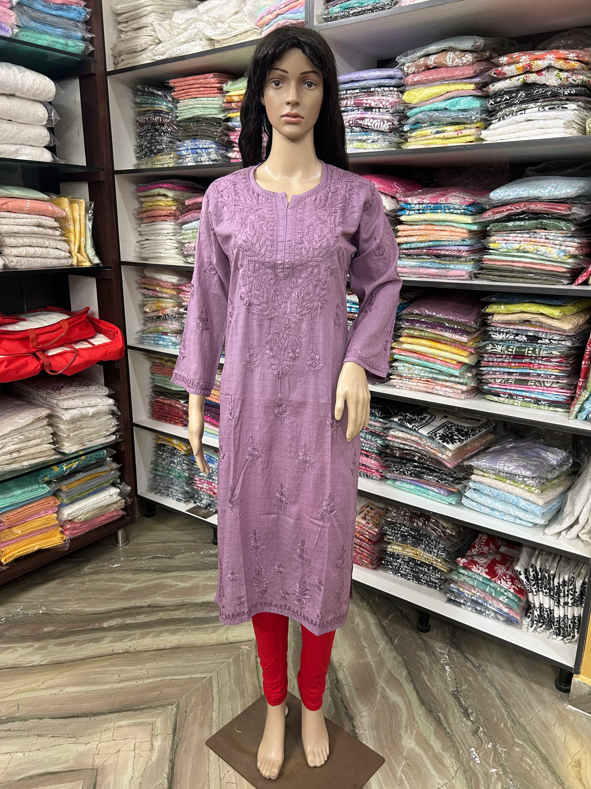 A mannequin wearing a lavender embroidered kurta with red leggings, displayed in a clothing store with shelves of folded garments in the background.