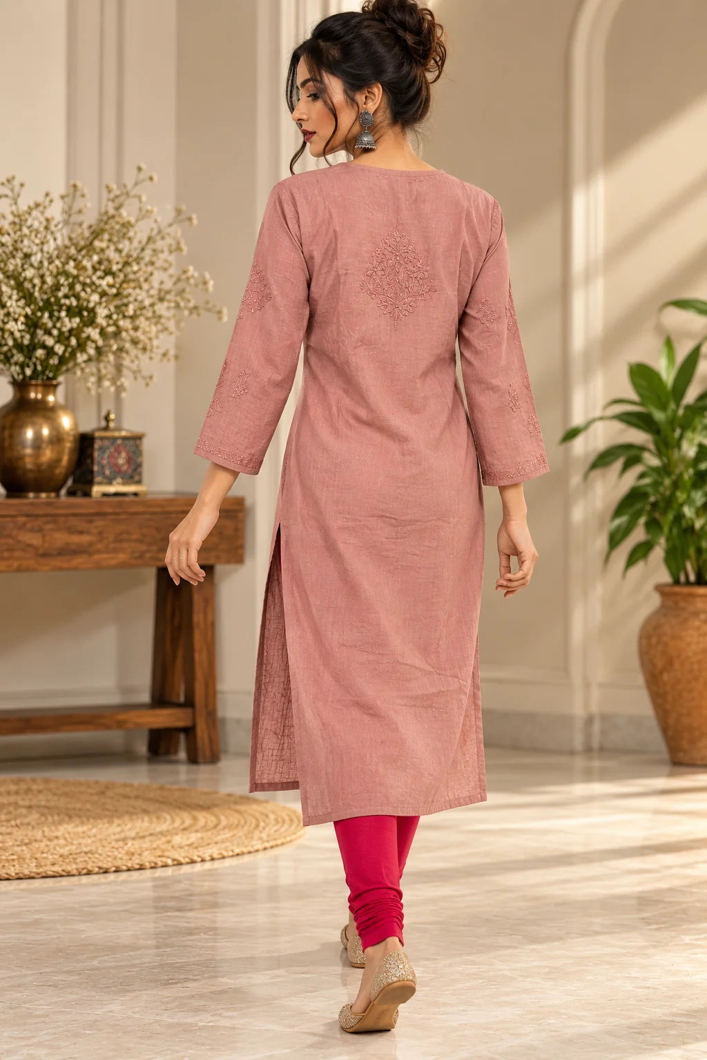 A woman wearing a dusty rose pink three-quarter sleeve kurta with subtle embroidery on the back, paired with bright pink leggings and beige flat shoes, shown from behind in a beautifully decorated indoor setting.