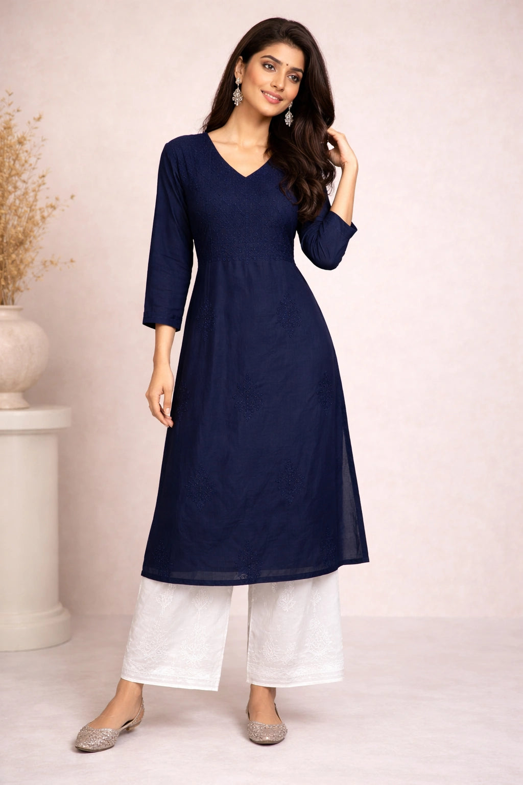 Lucknowi chikankari A woman modeling a navy blue V-neck kurta with three-quarter sleeves, paired with white wide-leg pants and silver flat shoes, standing against a light pink studio backdrop.