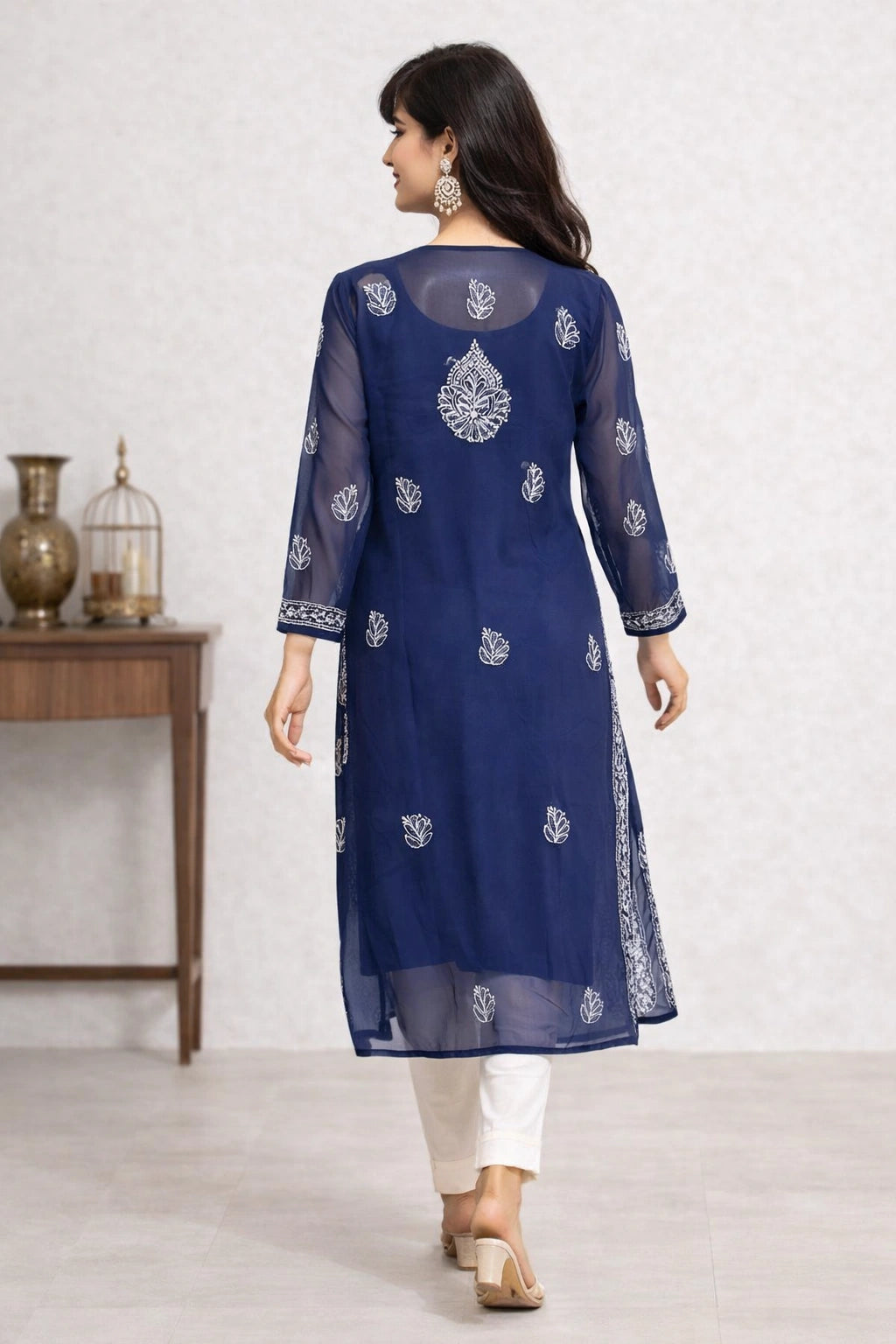 A woman wearing a navy blue sheer kurta with white embroidery, paired with white pants and beige sandals, shown from the back in an indoor setting.