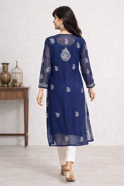 A woman wearing a navy blue sheer kurta with white embroidery, paired with white pants and beige sandals, shown from the back in an indoor setting.