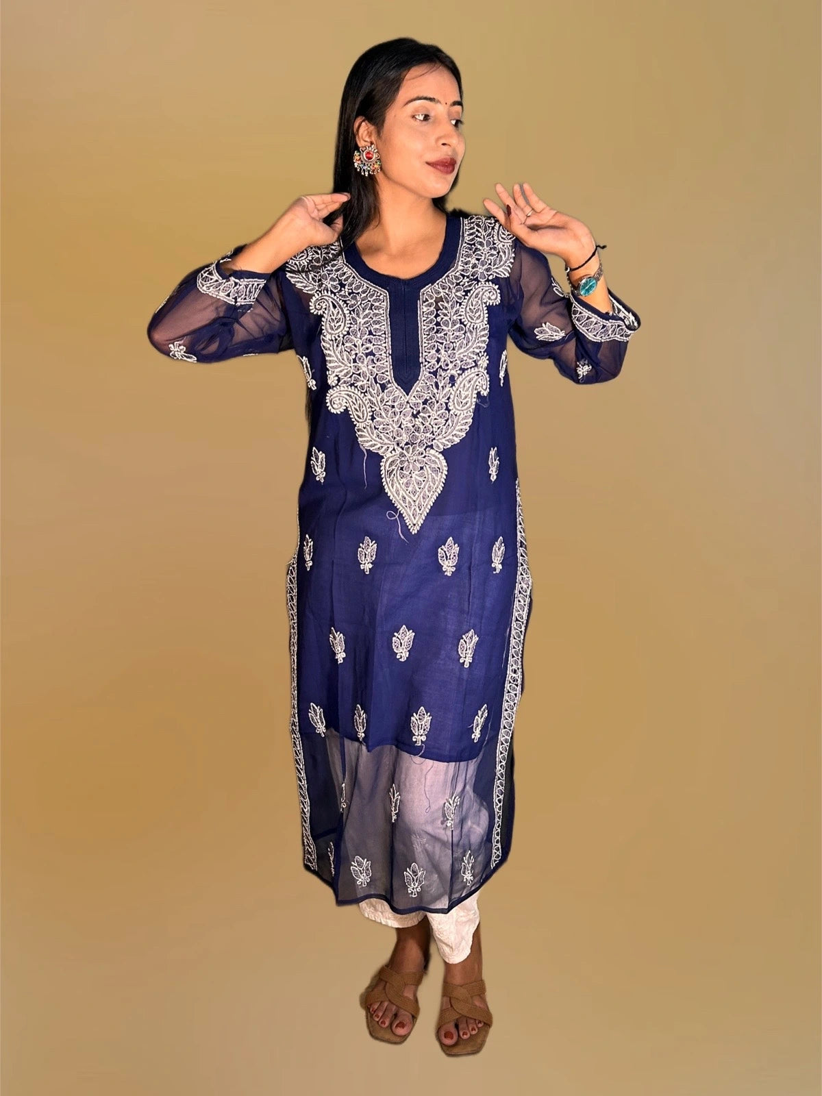 Lucknowi chikankari A woman modeling a navy blue embroidered kurta with white floral embroidery, paired with white pants and brown sandals, against a solid beige background.