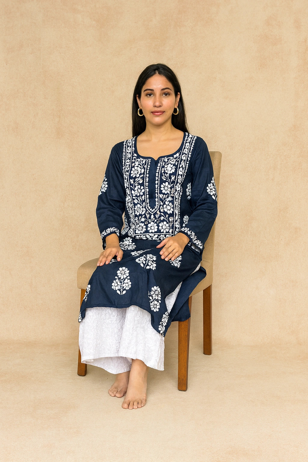 A woman sits on a wooden chair wearing a navy blue kurta with intricate white floral embroidery, paired with white trousers, against a textured beige backdrop.