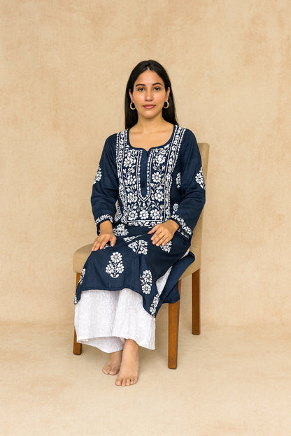 A woman sits on a wooden chair wearing a navy blue kurta with intricate white floral embroidery, paired with white trousers, against a textured beige backdrop.