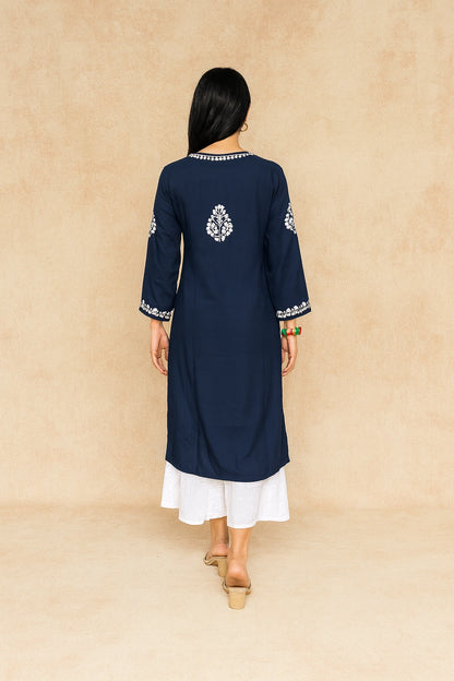 A woman viewed from the back wearing a navy blue kurta with white embroidered details on the shoulders, neckline, and cuffs, paired with a white skirt and beige sandals, set against a textured beige backdrop.