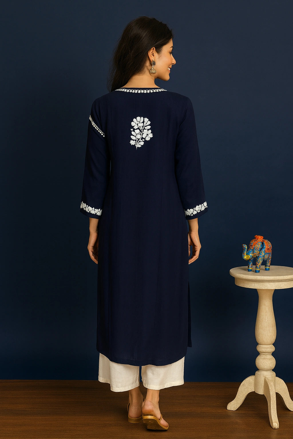 Woman wearing a navy blue embroidered kurta with white pants, standing against a dark blue wall.