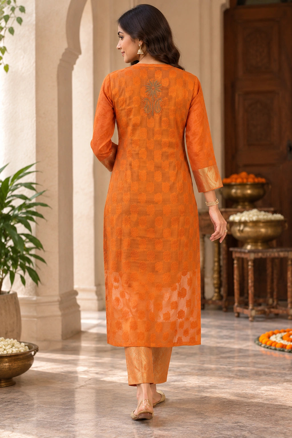 Lucknowi chikankari A woman wearing a bright orange kurta with matching trousers, photographed from behind in an elegant indoor courtyard setting with arches and decorative bowls.