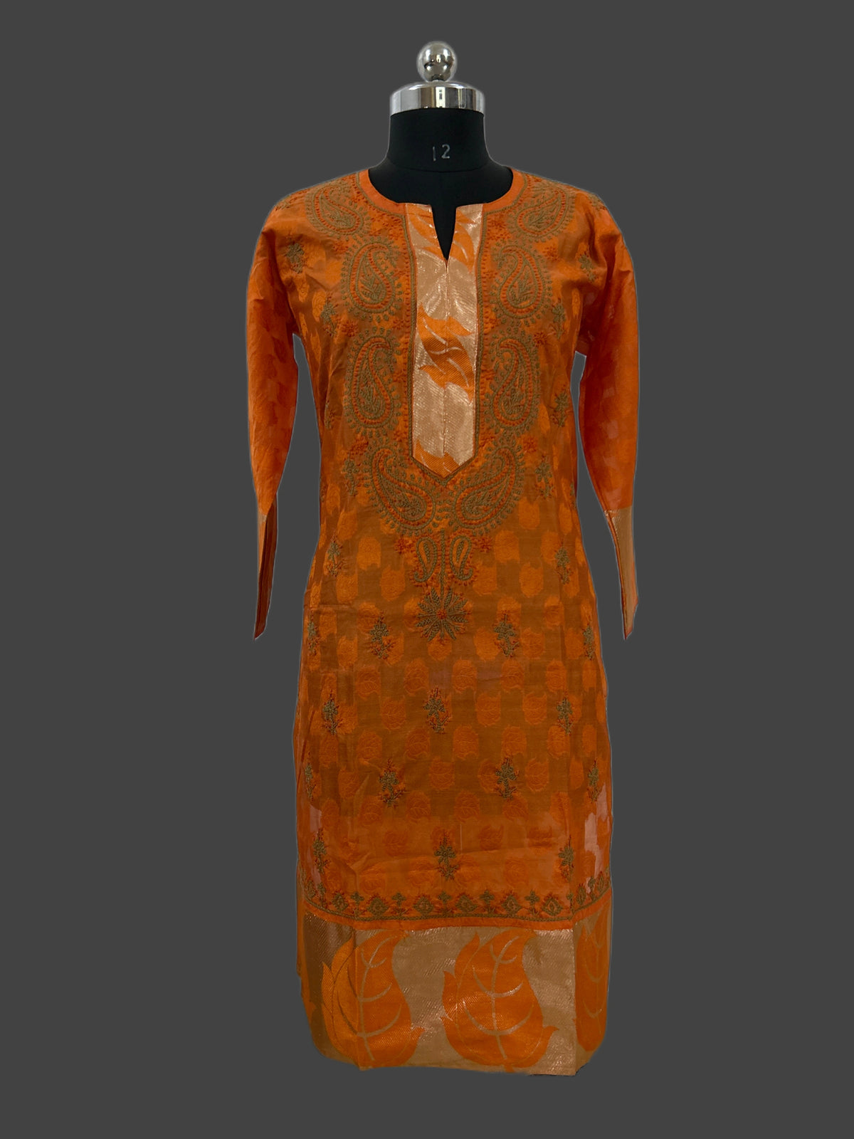 An orange embroidered kurta displayed on a black mannequin torso against a dark gray background. The kurta features a split neckline with a beige satin panel, intricate orange embroidery, and a matching orange and beige patterned hem.