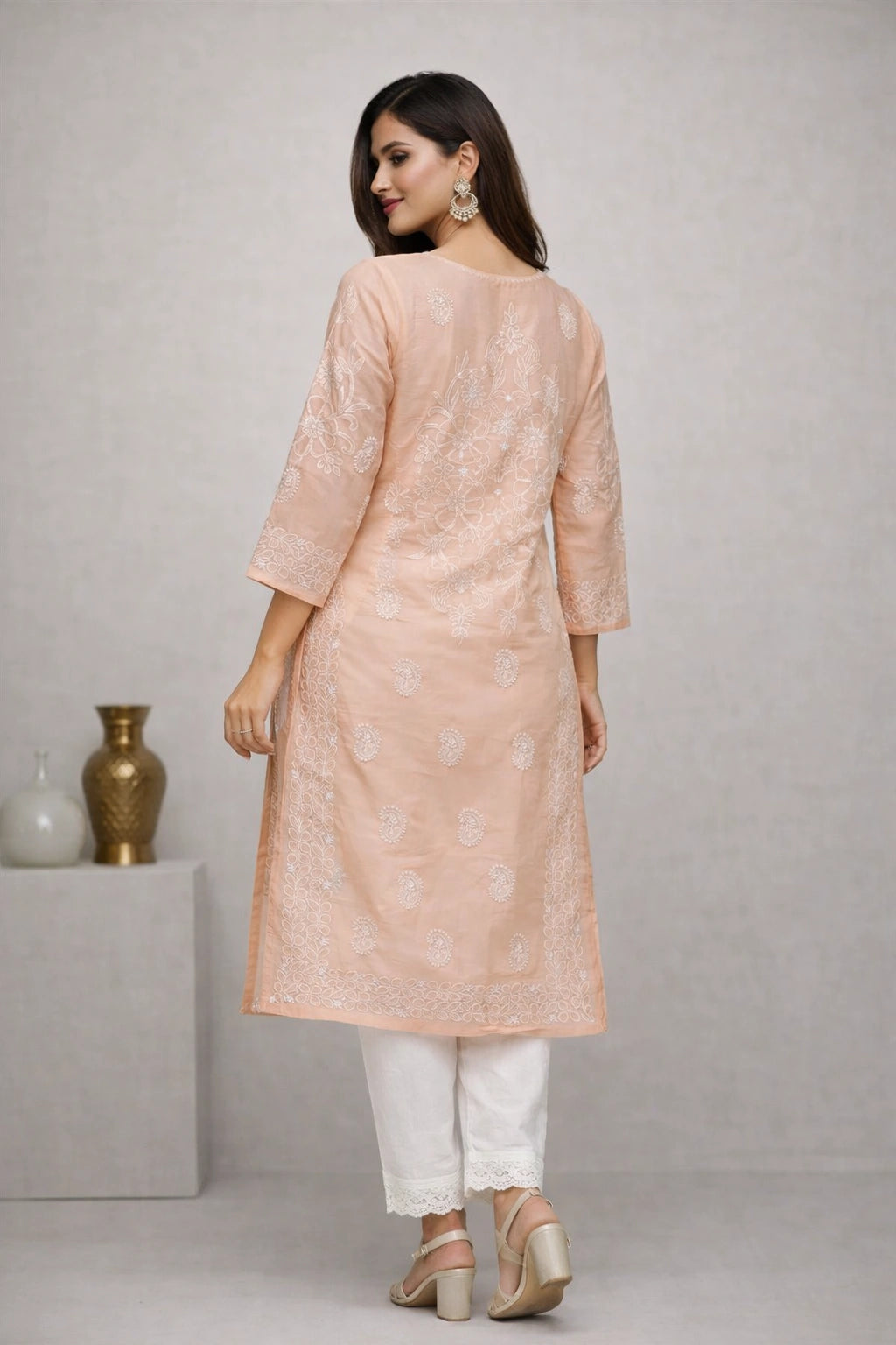 A woman modeling a peach-colored embroidered kurta with white pants and beige heels, shown from the back against a neutral gray studio background.