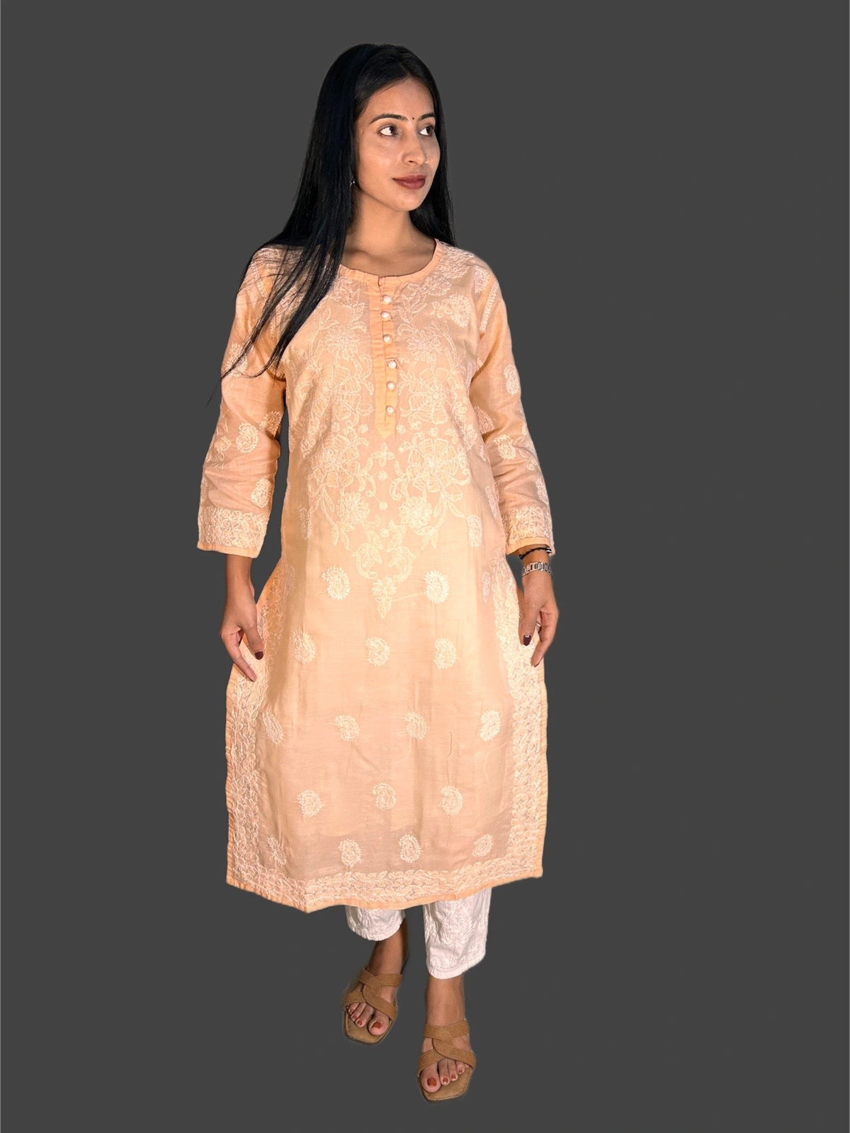 A woman modeling a peach-colored embroidered kurta with white pants and brown sandals, set against a solid dark gray background.