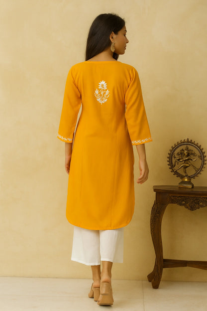 Woman wearing a yellow kurta with white embroidery, standing against a beige wall.