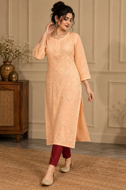 A woman models a peach-colored embroidered kurta with three-quarter sleeves, paired with dark red pants and beige shoes, standing in a decorated room with a wooden cabinet and neutral wall paneling.