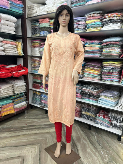 A peach-colored embroidered kurta displayed on a female mannequin, paired with red leggings, set against a background of clothing racks filled with folded garments.