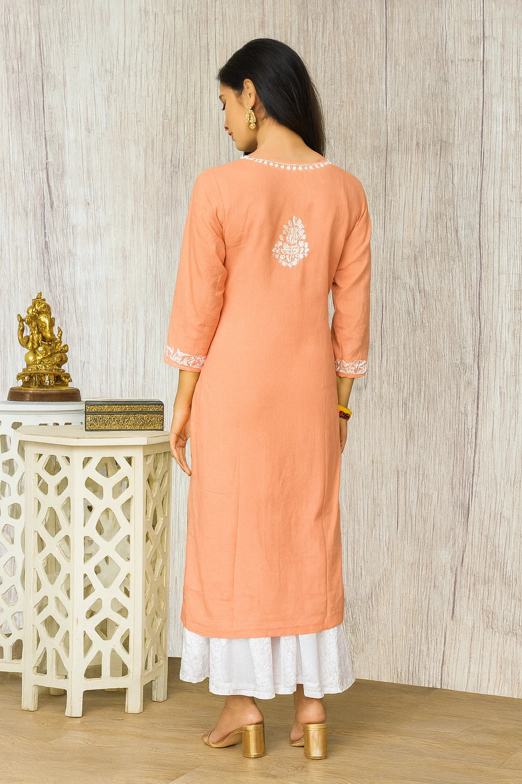 A woman is shown from the back wearing a peach-colored kurta with white embroidery on the back and cuffs, paired with a white flared skirt. She is standing in front of a light wood-paneled wall next to a decorative white side table.