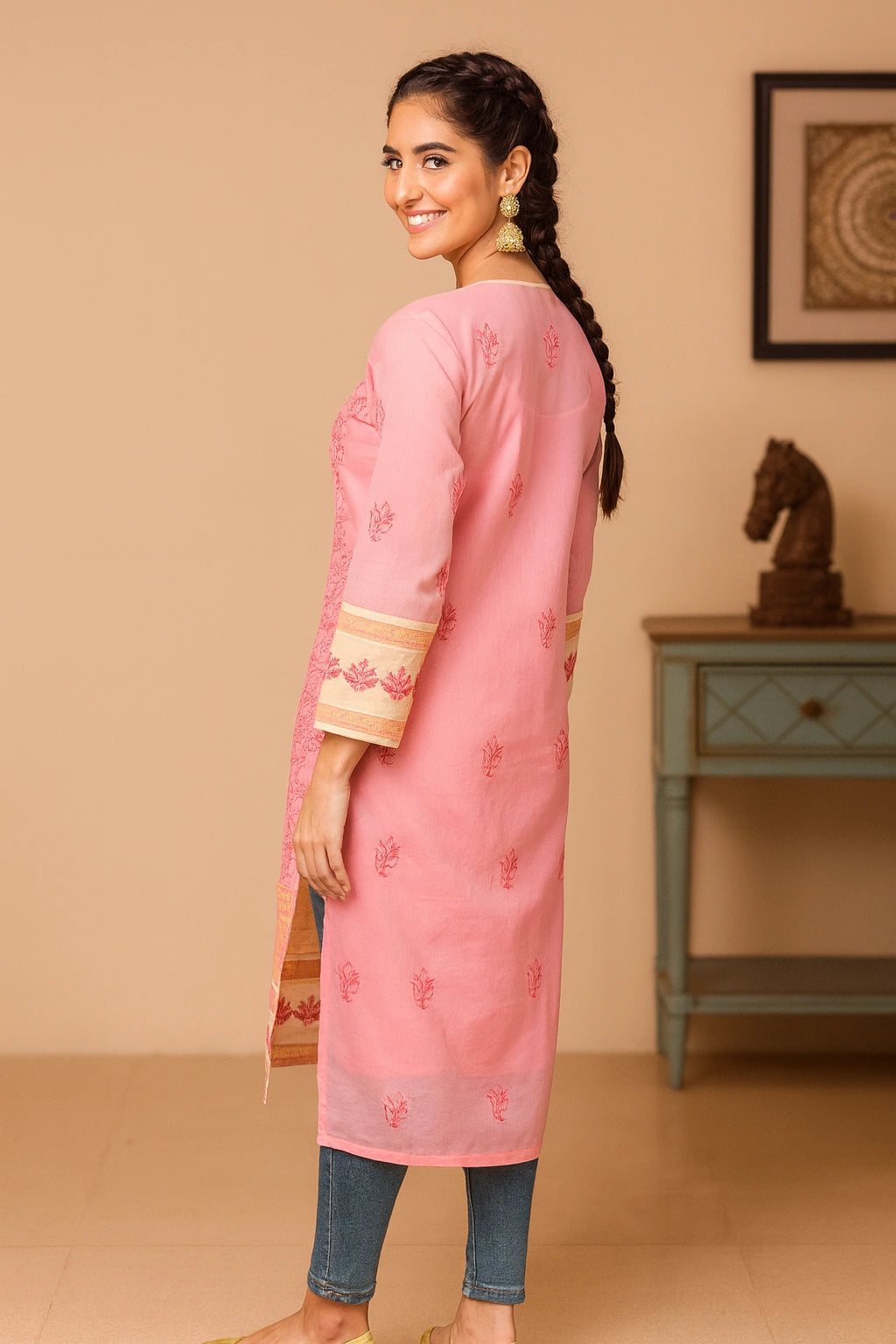 A woman viewed from behind wearing a pink long kurta with three-quarter sleeves and a subtle embroidered motif on the back, paired with denim and beige flat shoes in an indoor setting.
