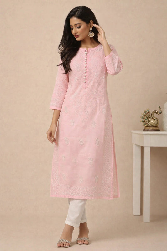 A woman modeling a light pink embroidered kurta with white pants, standing in front of a beige wall next to a small white table with a decorative peacock figurine.