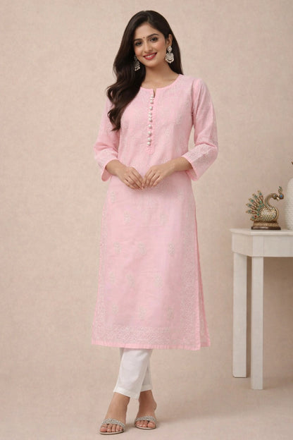 lucknowi chikankari A woman modeling a light pink long kurta with white pants, standing against a beige studio backdrop with a small white side table and decorative peacock figurine in the background.