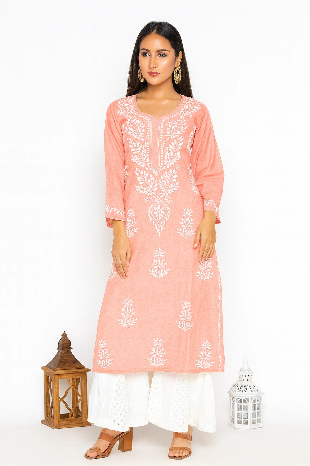 A woman modeling a peach-pink long kurta with white floral embroidery, paired with white palazzo pants, standing against a white studio background with decorative lanterns.