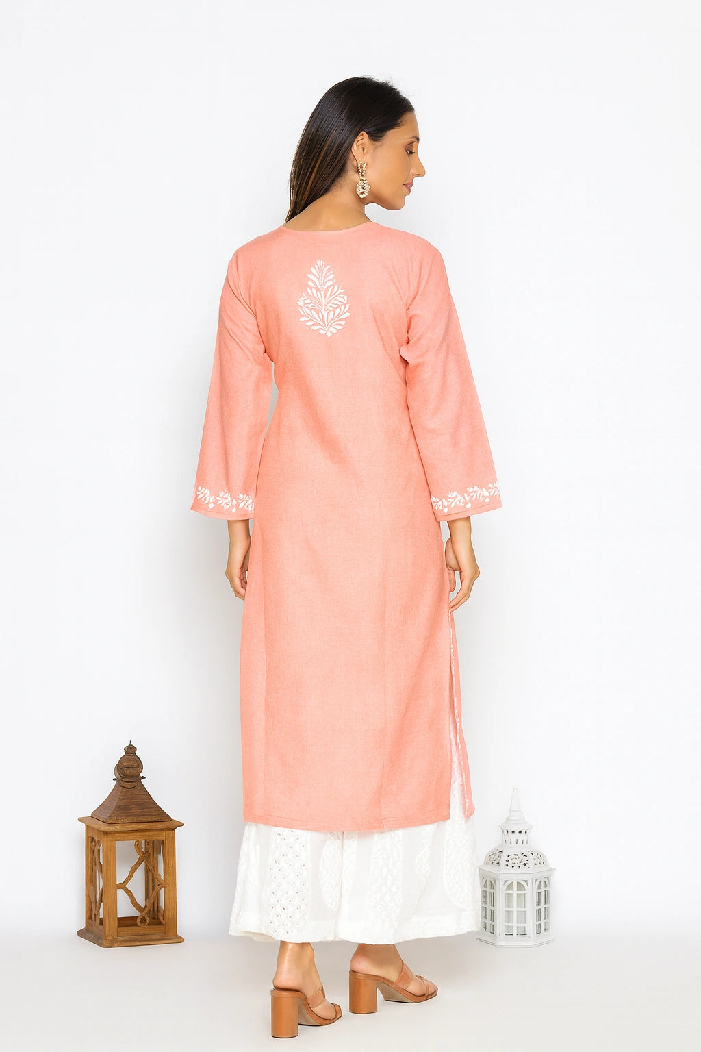 A woman is shown from the back wearing a peach-pink long kurta with white embroidery on the back and sleeve cuffs, paired with a white skirt. She is standing against a white studio background with decorative lanterns on the floor.