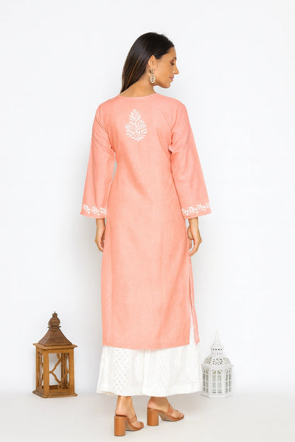 A woman is shown from the back wearing a peach-pink long kurta with white embroidery on the back and sleeve cuffs, paired with a white skirt. She is standing against a white studio background with decorative lanterns on the floor.