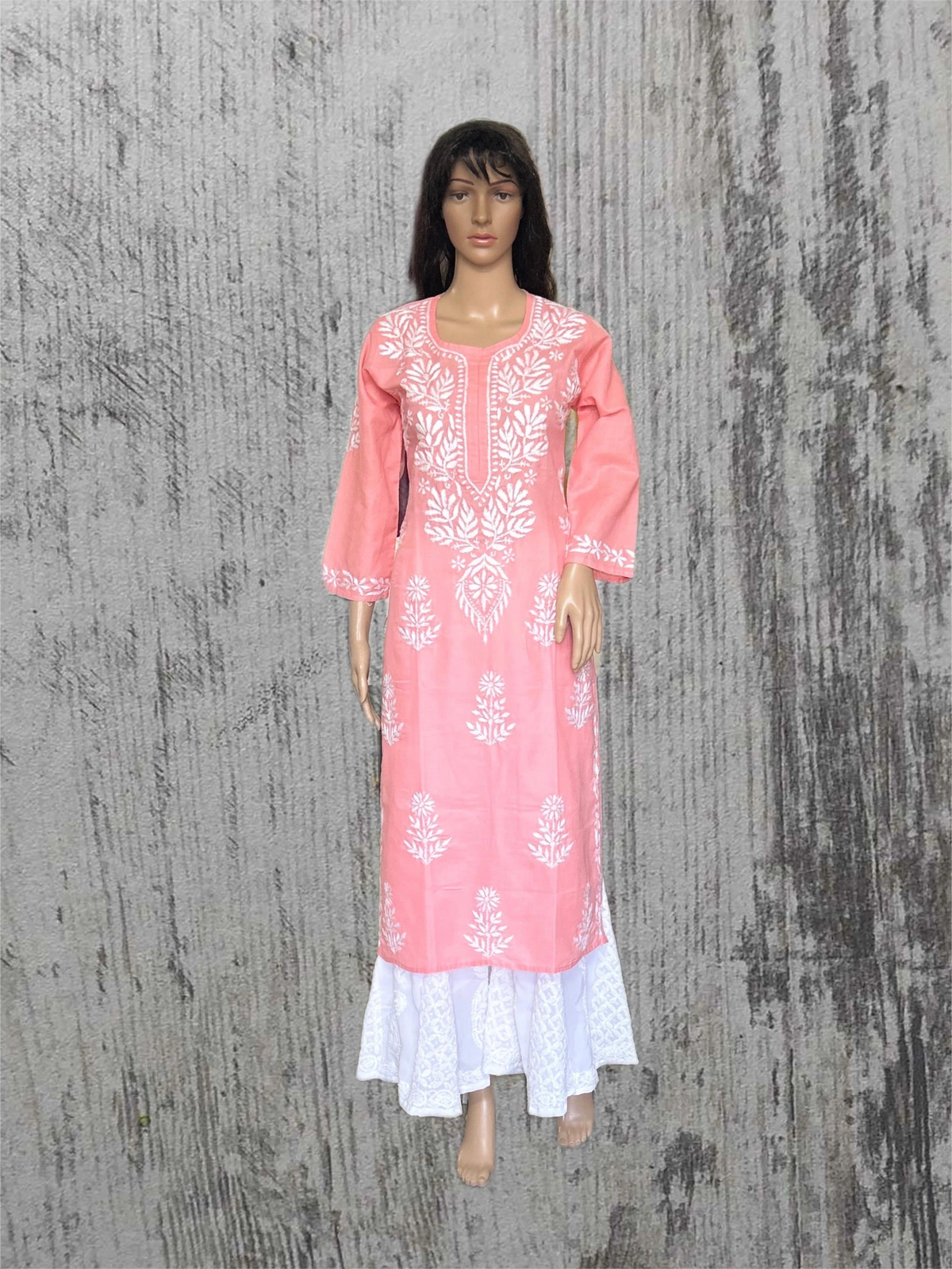 A pink long kurta dress with white floral embroidery and a white ruffled hem, displayed on a female mannequin against a textured gray background.