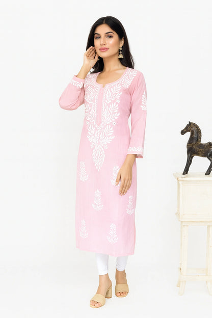 A woman modeling a light pink long kurta with white floral embroidery, paired with white leggings and beige sandals, standing against a white studio background next to a small table with a horse figurine.