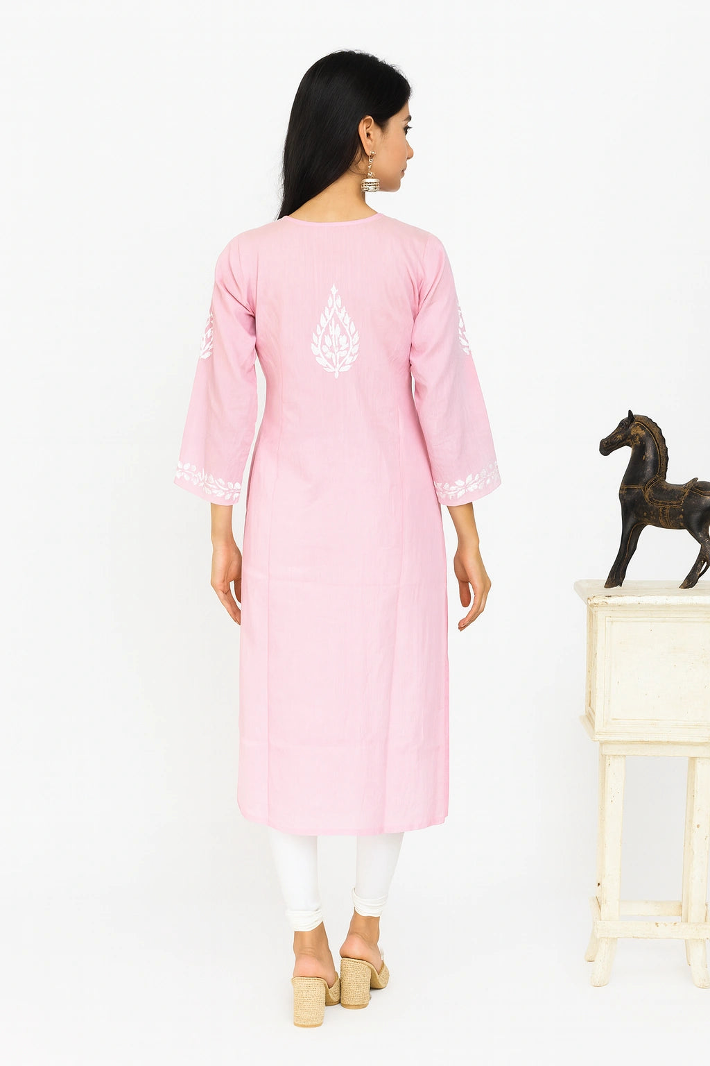 A woman viewed from the back wearing a light pink tunic with white embroidered motifs on the back and sleeve cuffs, paired with white leggings and beige block-heel sandals, set against a plain white studio background with a small decorative horse statue on a side table.