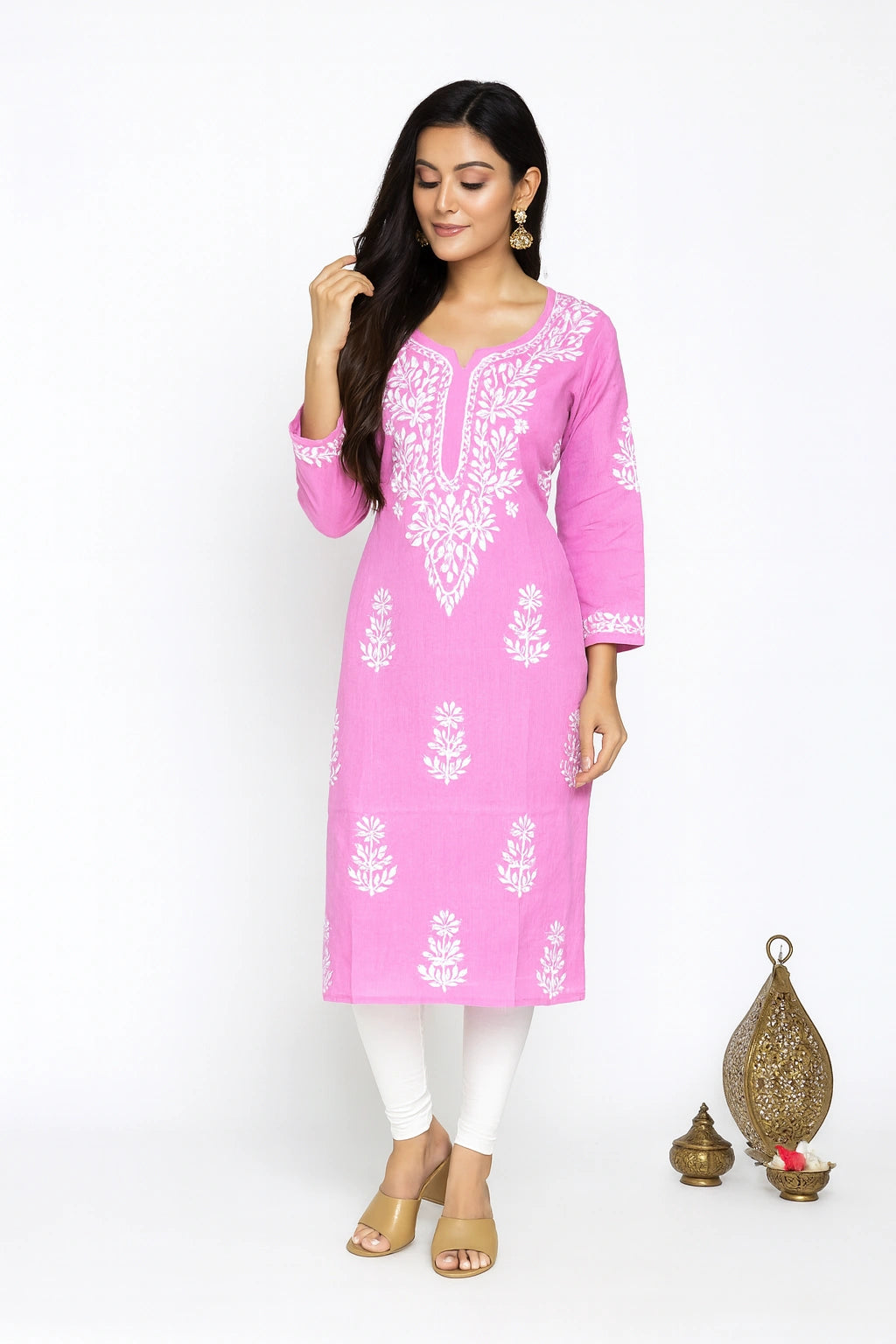 A woman modeling a pink long kurta with white floral embroidery, paired with white leggings and beige sandals, standing against a white studio background with decorative brass lamps on the right.