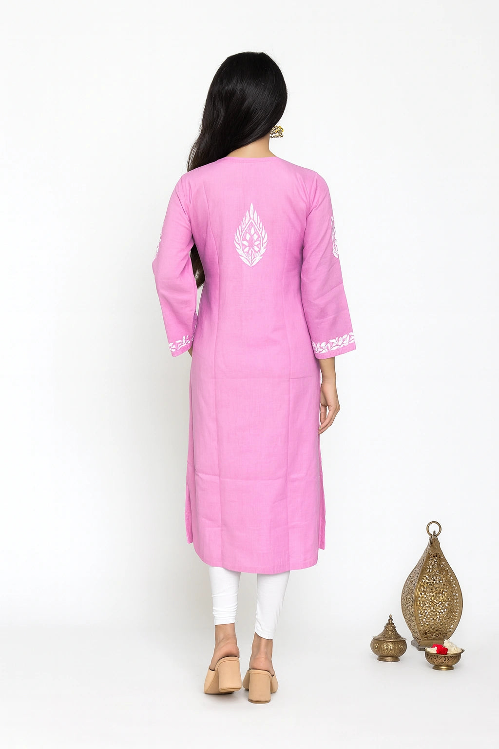 A woman viewed from the back wearing a pink kurta with white embroidered details on the back and cuffs, paired with white leggings and beige flat shoes, set against a plain white background with decorative brass vessels on the right.