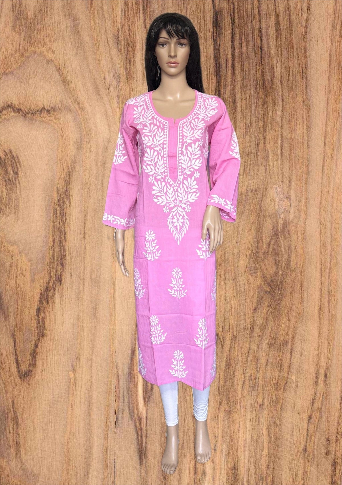 A pink long kurta with white floral embroidery, displayed on a female mannequin against a brown wood-grain background.