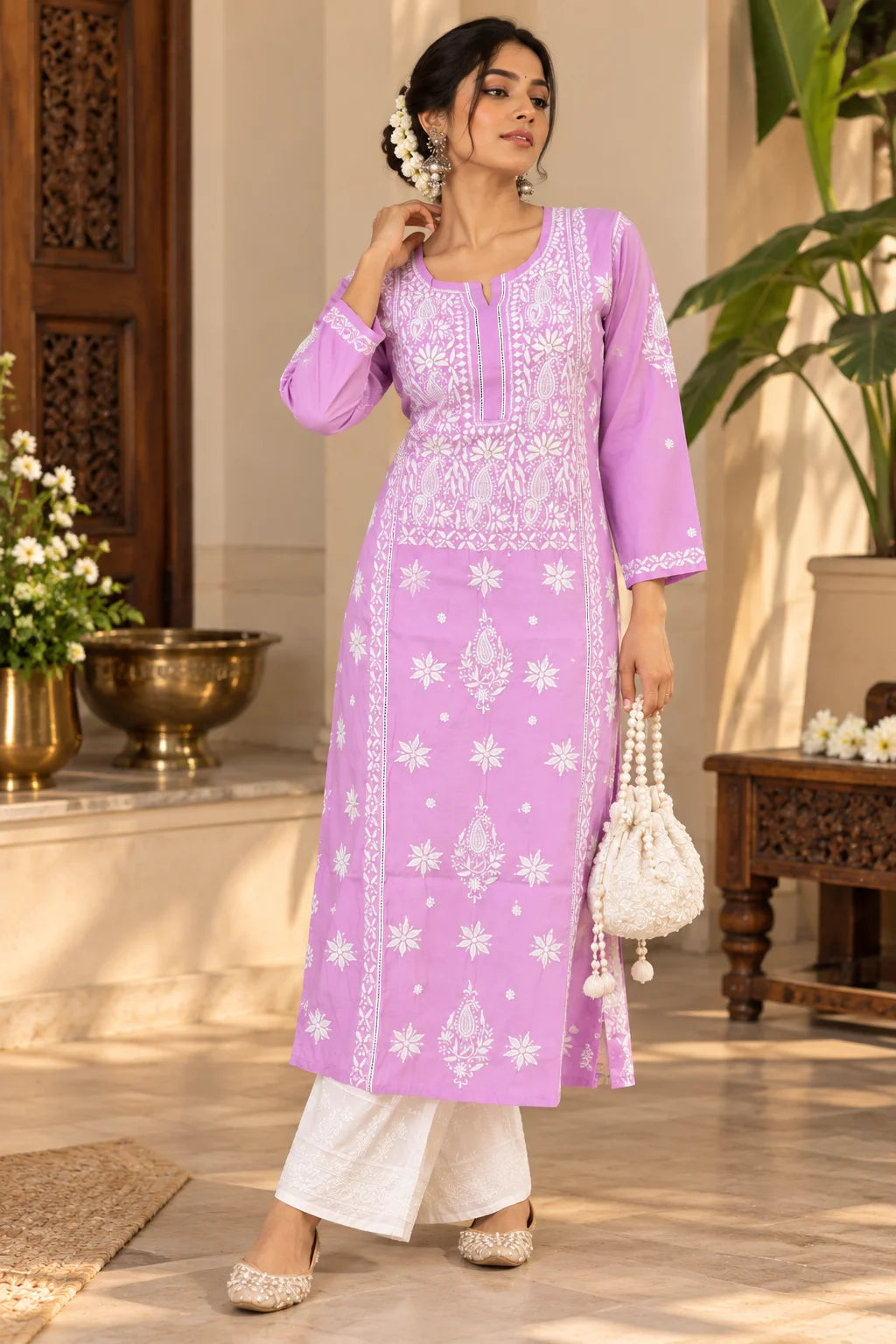 A woman models a light pink long kurta with white floral embroidery and white pants in a warm, decorated indoor setting.