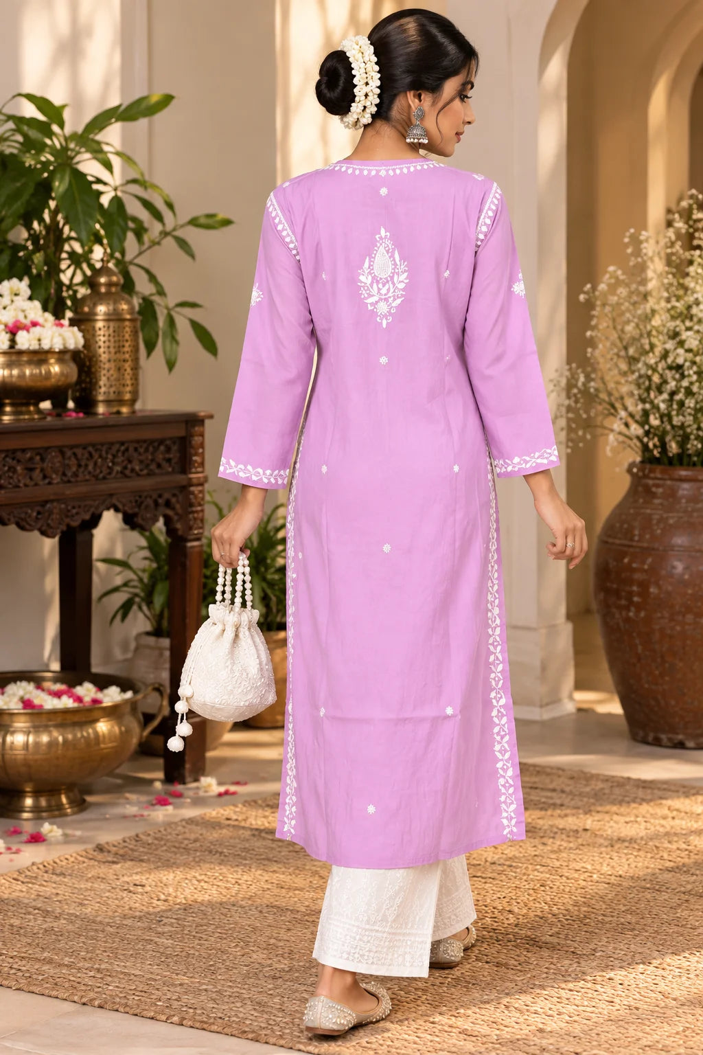 A woman wearing a light pink long kurta with white embroidered details on the back, paired with white pants, walking in a warm, sunlit indoor setting.